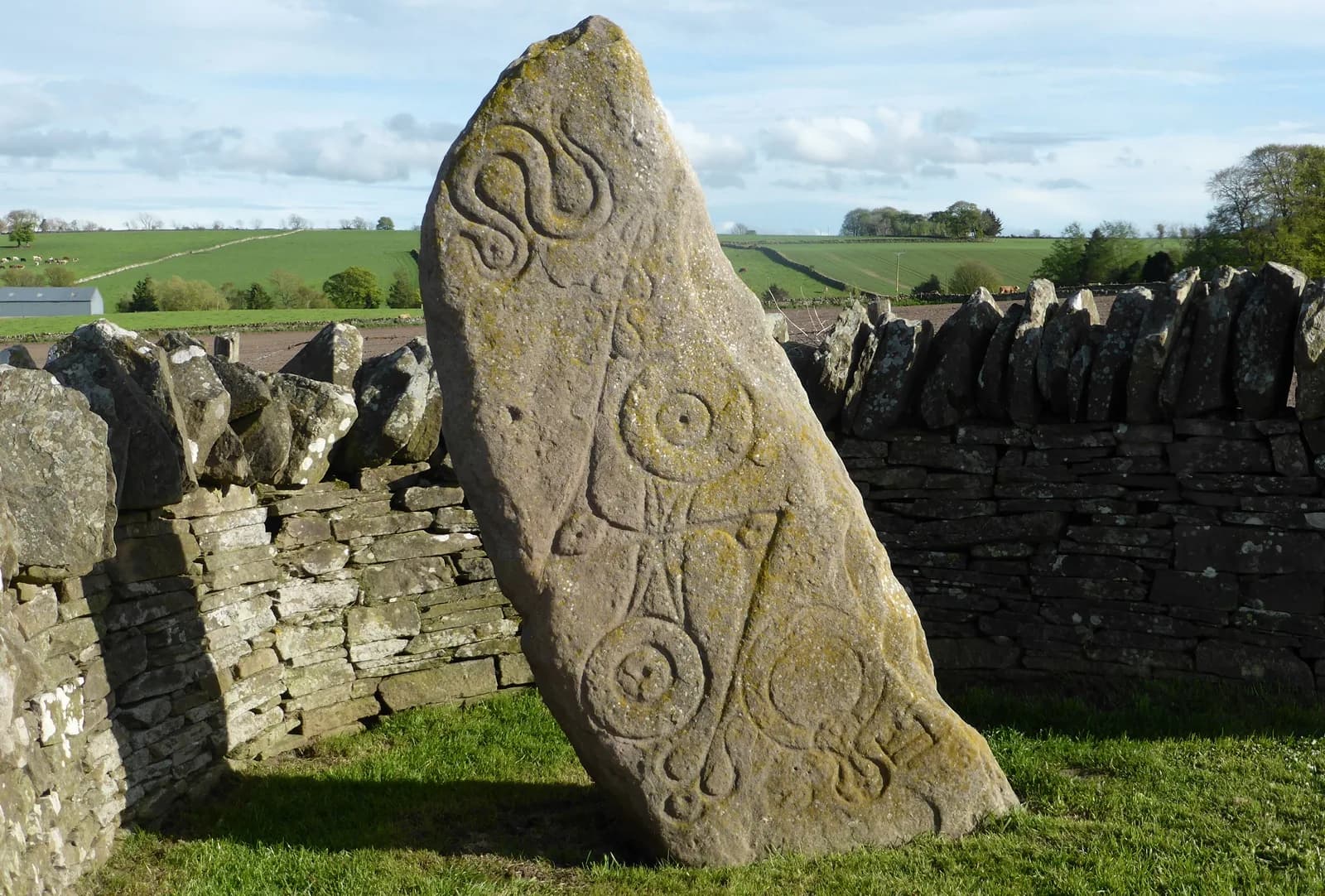 Weathered carved upright stone beside a grassy roadside.