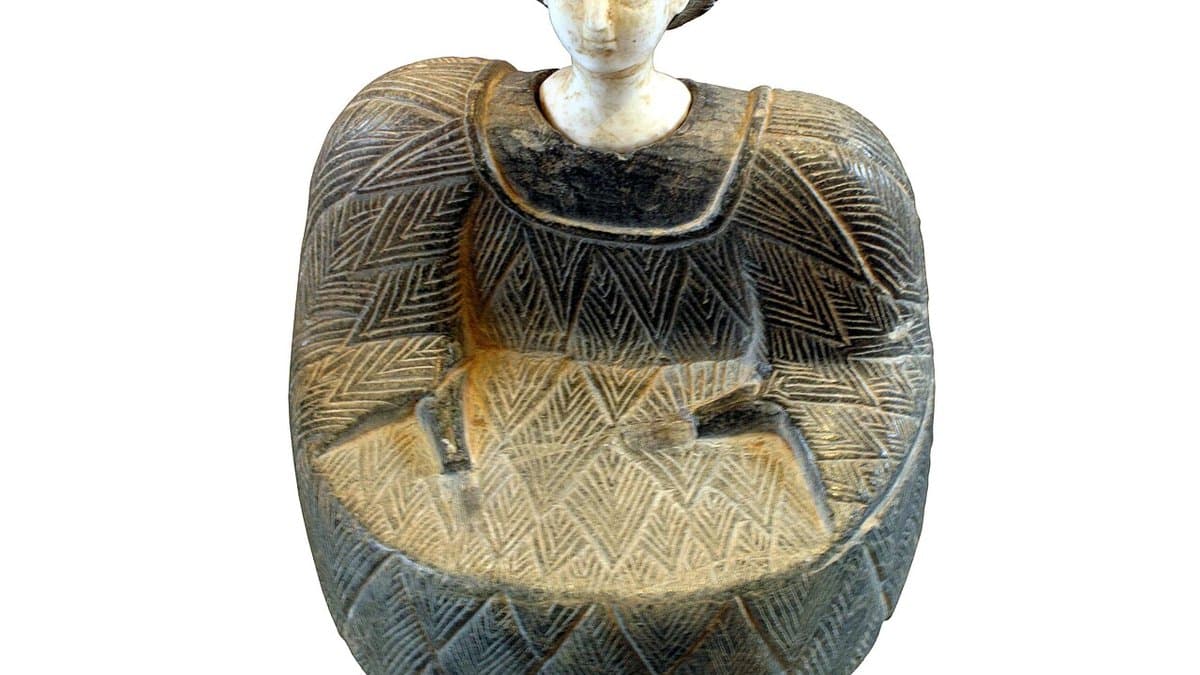 Seated Bactrian figurine with a pale stone head and a dark garment carved with a herringbone pattern.
